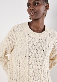 Flauschiger, creamfarbener Strickpullover mit einer Mischung aus strukturierten Rauten- und offenen Maschen. Er hat einen weiten gerippten Halsausschnitt und eine lockere Passform.