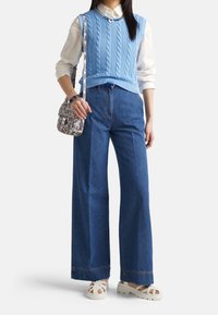 Suéter sin mangas de punto con cable en azul claro sobre una camisa blanca, combinado con jeans azules de pierna ancha de talle alto y sandalias blancas de plataforma.