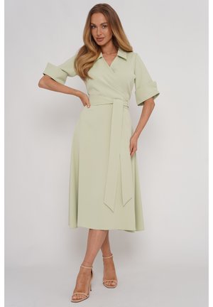 Kobieta nosząca jasnobeżową sukienkę midi wiązaną, z podwiniętymi rękawami i długim paskiem w talii, w połączeniu z beżowymi szpilkami na pasku wokół kostki, stojąca na białym tle.