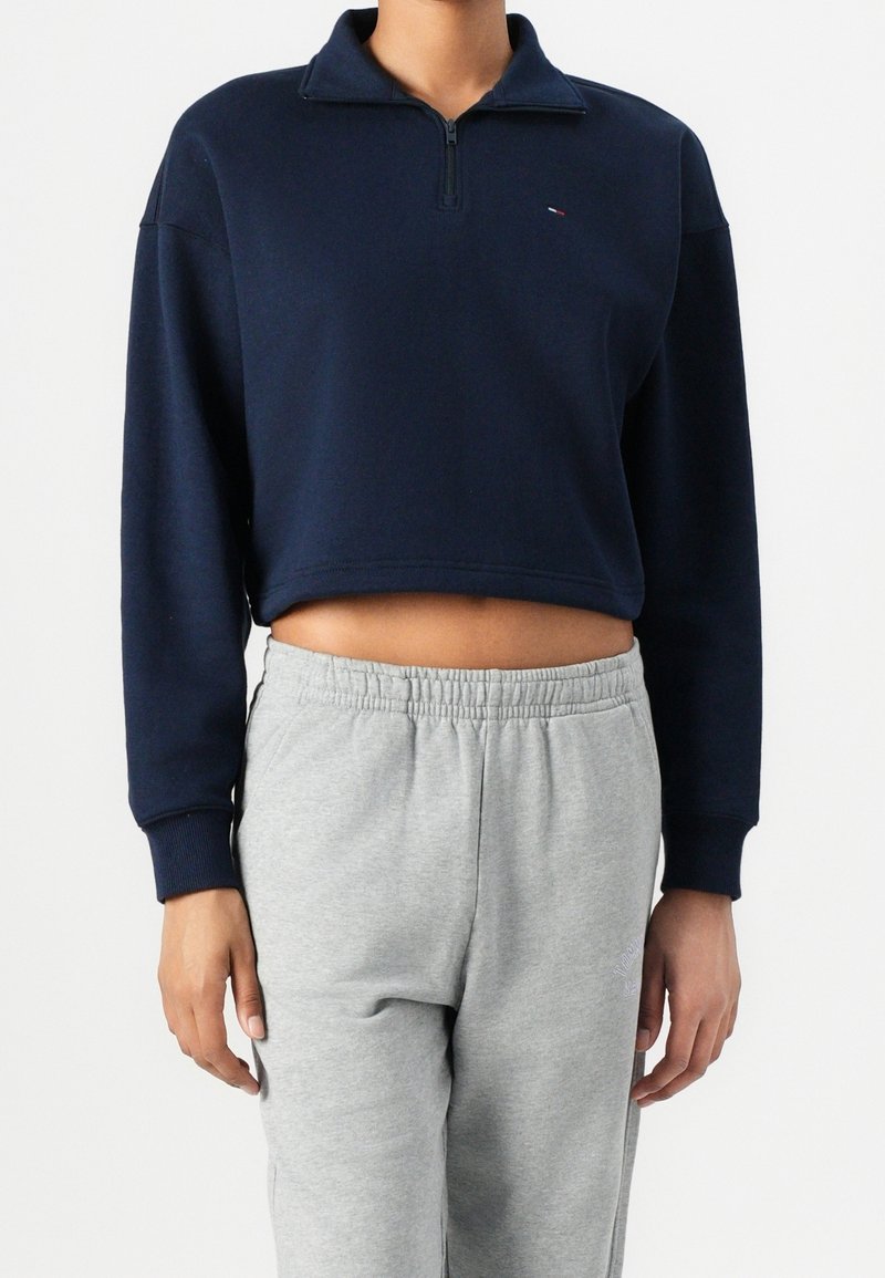 Personne portant un sweat-shirt court bleu marine à fermeture éclair quart de zip et un pantalon de jogging gris clair à taille élastique sur un fond uni.