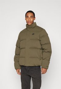 Giacca puffer verde oliva con colletto alto, dotata di chiusura con zip e logo Nike nero sul petto. Texture morbida e forma squadrata.