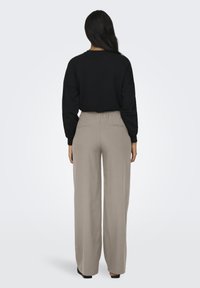 Donna con lunghi capelli scuri, vestita con un top nero a maniche lunghe e pantaloni beige a gamba larga, in piedi di spalle su uno sfondo neutro.