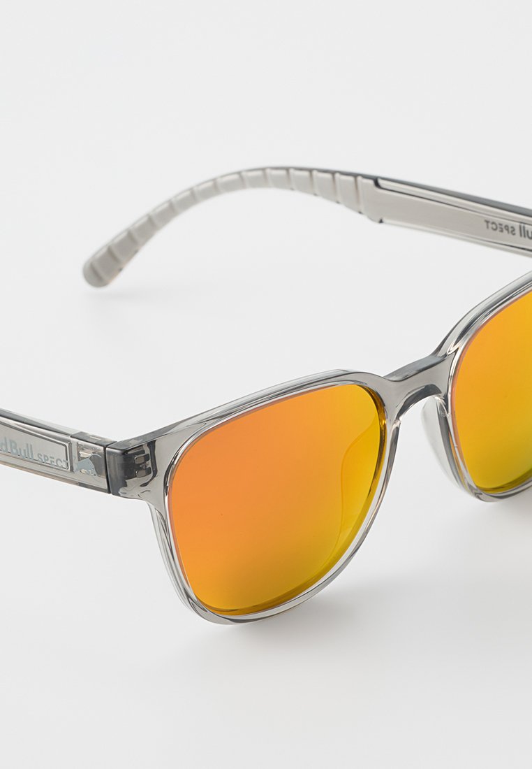 Okulary przeciwsłoneczne z przezroczystą szarością, pomarańczowymi, lustrzanymi soczewkami i teksturowanymi szarymi zausznikami z detalem marki po boku.
