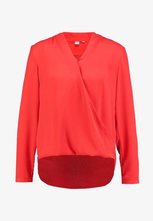 Blouse portefeuille rouge avec un col en V, des manches longues, et une texture douce et lisse. Associée à un jean foncé, avec peu de détails supplémentaires.