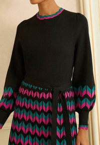 Rochie neagră cu partea superioară mulată, mâneci bufante și un model în zigzag multicolor în tonuri de teal și roz pe fustă. Dispune de o talie legată și o decolteu cu margini ondulate.