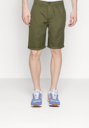 Pantalones cortos de algodón verde oliva con dobladillos enrollados, con cierre de botón. Llevados con zapatillas azules y naranjas con suelas texturizadas.