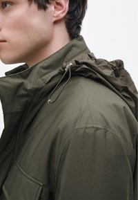 Olivengrøn jakke med høj krave, lynlåslomme og justerbar snøre i hætten. Glat tekstur, struktureret design og minimal detaljering.