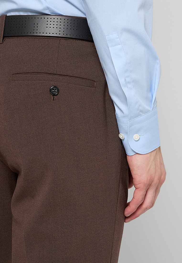 Pantaloni marroni con una texture liscia, dotati di una tasca posteriore chiusa con un bottone nero. Una camicia azzurro chiaro è parzialmente visibile.