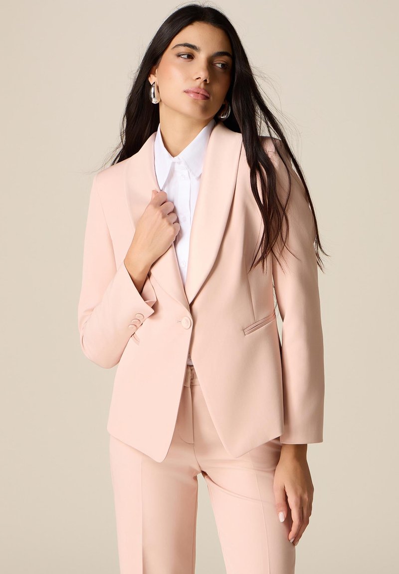 Frau mit langen dunklen Haaren, die ein hellrosa maßgeschneidertes Blazer und passende Hose, ein weißes Hemd mit Kragen und silberne Creolen trägt, seitlich blickend.