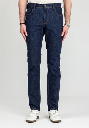 Mężczyzna ubrany w ciemnoniebieskie, dopasowane dżinsy i białe trampki stoi na tle jednolitego jasnego tła.