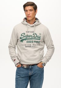 Sudadera gris con cordones y un bolsillo frontal. Texto del logo verde en el pecho. Camisa de cuadros visible en el cuello. Llevada con jeans azules.
