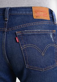 Donkerblauwe spijkerbroek van denim met een bruine leren logopatch, oranje stiksel en een kenmerkend gebogen zakdesign.