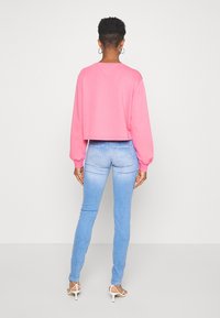 Mujer de pie mirando hacia atrás, vestida con un sudadera corta rosa, jeans ajustados azul claro, sandalias de tacón blancas y pendientes de aro plateados.