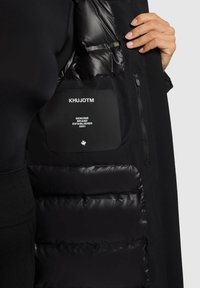 Veste rembourrée noire avec un matériau extérieur lisse. Comprend une étiquette noire affichant le nom de la marque et les détails, ainsi qu'une poche zippée à l'intérieur.
