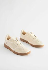 Beige sneakers with a textured upper, lace-up design, and a gum rubber sole. Features include stitched accents and perforated areas on the toe.