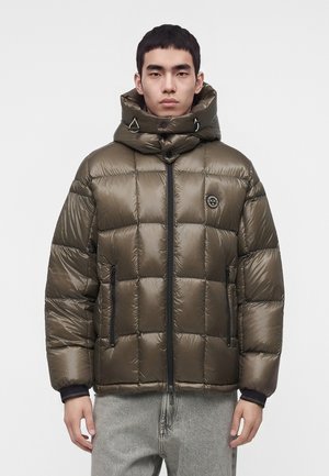 Homme portant une doudoune vert olive brillante avec un col montant, une fermeture éclair frontale et des poches latérales zippées, associée à un jean gris clair.