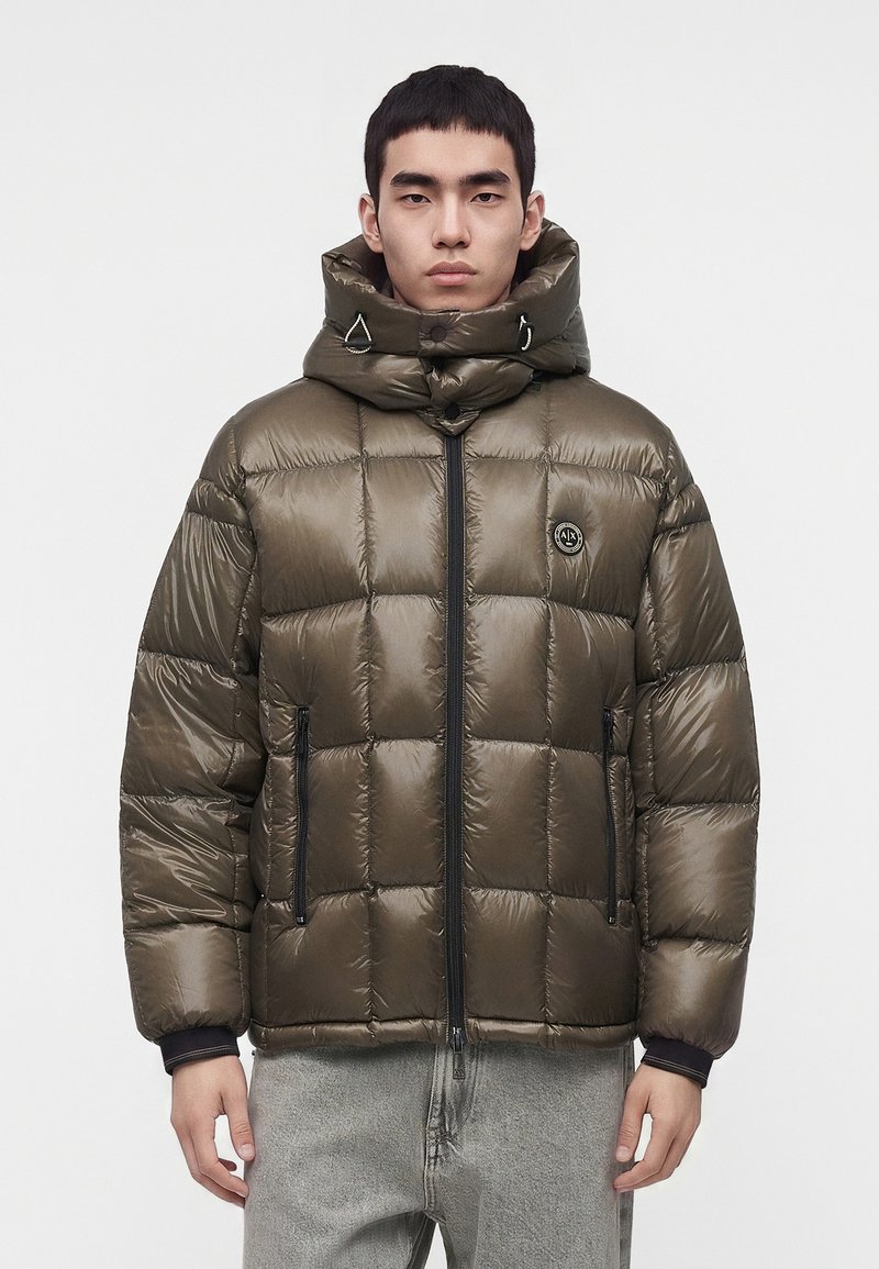 Homme portant une doudoune vert olive brillante avec un col montant, une fermeture éclair frontale et des poches latérales zippées, associée à un jean gris clair.