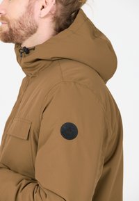 Brun huva jacka gjord av slät tyg, med en patch på ärmen, framficka och justerbar dragsko vid huvudet.