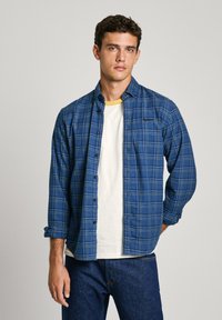 Camicetta di flanella a quadri blu con bottoni sul davanti, maniche arrotolate e una t-shirt bianca a collo rotondo sotto, abbinata a jeans in denim scuro.