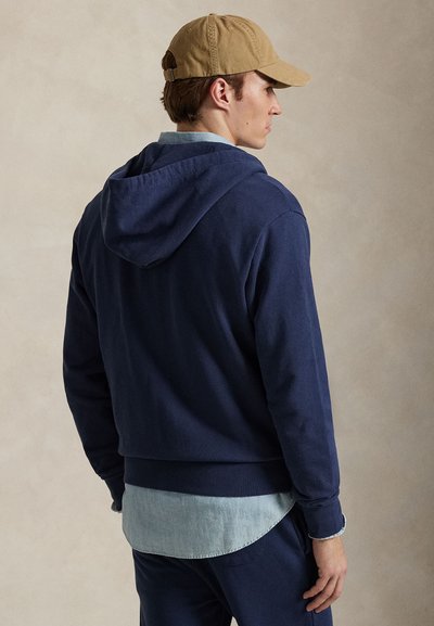 Tmavomodrá mikina s kapucňou na sťahovanie, mäkkým materiálom a rebrovanými manžetami. Nosená cez svetlomodrú denimovú košeľu. Ležérny strih.