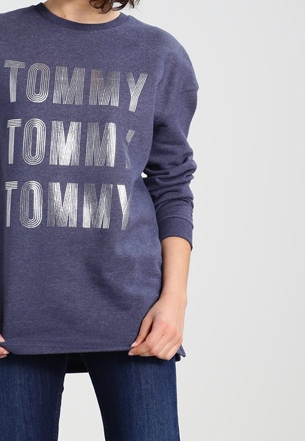 Felpa blu navy con grande scritta bianca "TOMMY" a righe, scollo tondo e maniche lunghe, abbinata a jeans.