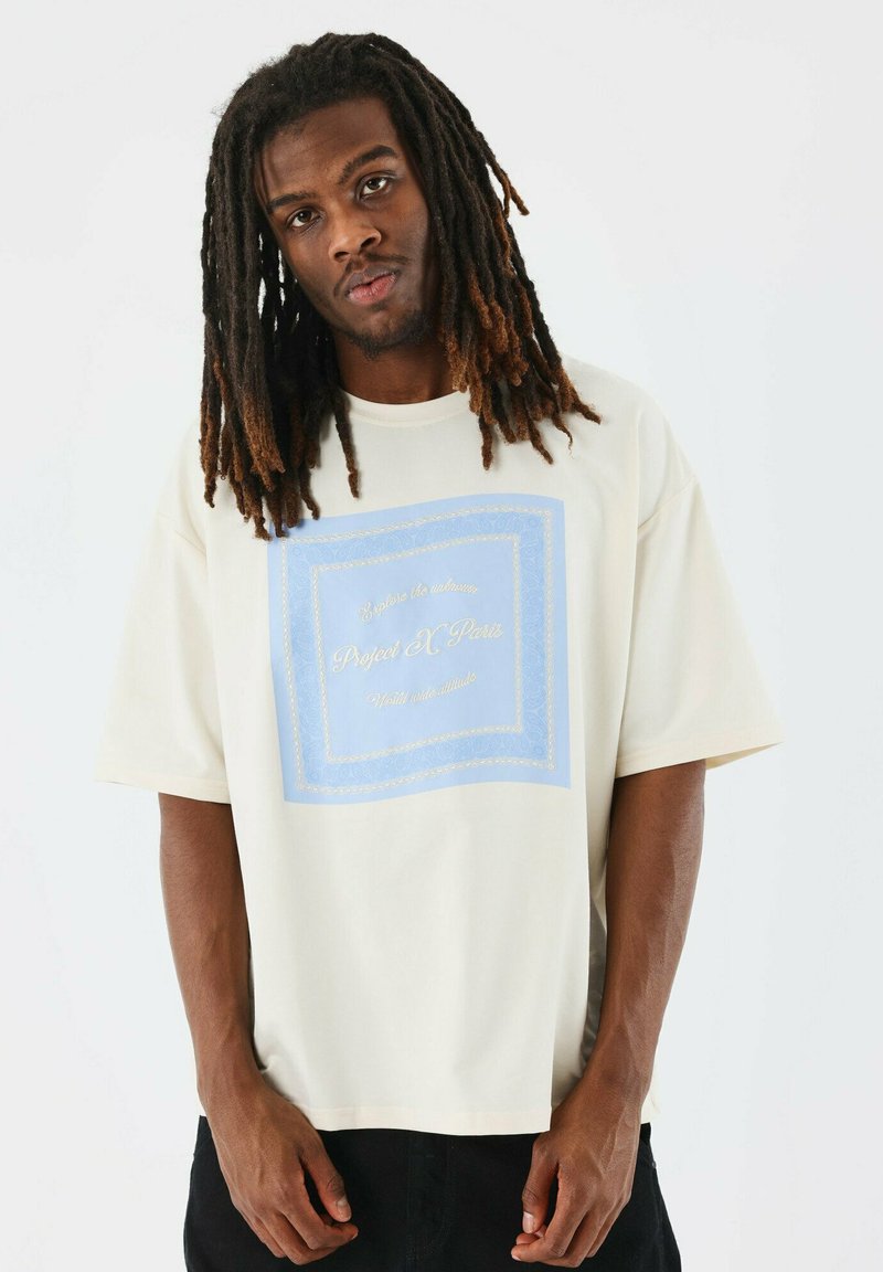 Camiseta de gran tamaño color crema, con un gráfico rectangular azul claro con texto dorado en el centro. Manga corta, tejido suave.