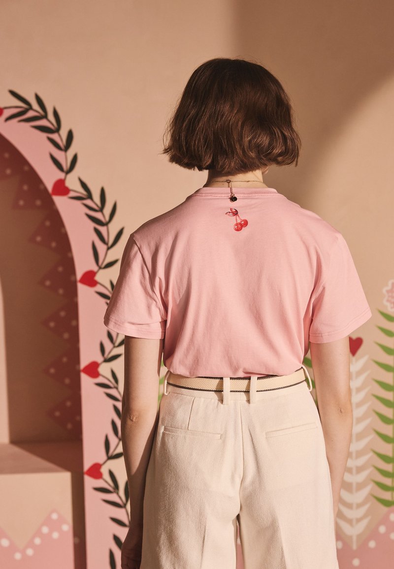 Růžové bavlněné tričko s krátkými rukávy, na zádech s červeným motivem třešní, doplněné béžovými kalhotami s vysokým pasem a černým pásem.
