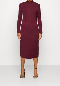 Kvinna som bär en långärmad, ribbad, vinröd midiklänning tillsammans med svarta högklackade sandaletter med ankelrem, stående mot en enfärgad bakgrund.