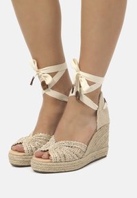 Sandales compensées beige en espadrille avec une tige tissée, bout ouvert et liens à la cheville. Talon recouvert de corde et texture à carreaux à l'arrière.