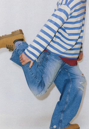 Osoba koja nosi smeđe čizme, poderane plave traperice i plavo-bijeli prugasti džemper savija jedno koljeno i drži stopalo iza sebe.