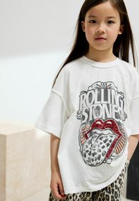 Biała koszulka z krótkim rękawem z logo Rolling Stones z czerwonymi cekinowymi ustami i językiem w zwierzęcy wzór, noszona z spodniami w panterkę.