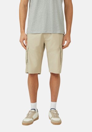Mann trägt beige Cargoshorts, weiße Socken und beige Sneaker mit weißen und dunkelblauen Details, steht vor einem schlichten Hintergrund.