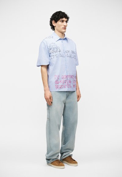 Jeune homme aux cheveux bouclés portant une chemise à manches courtes rayée bleue avec du texte, un jean clair ample et des baskets marron, debout devant un fond blanc.