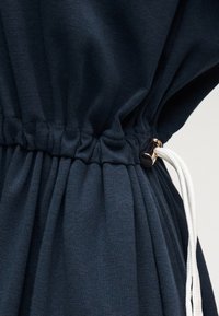 Gros plan sur une taille en tissu bleu foncé froncé avec un cordon blanc réglable et un curseur en plastique noir.