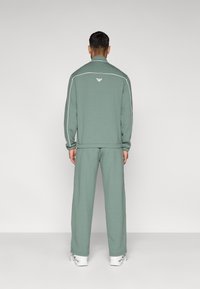 Πράσινο φόρμα από μαλακό ύφασμα, με ζακέτα με φερμουάρ και λευκές ρίγες, καθώς και λογότυπο στην πλάτη, συνδυασμένη με φαρδύ παντελόνι.