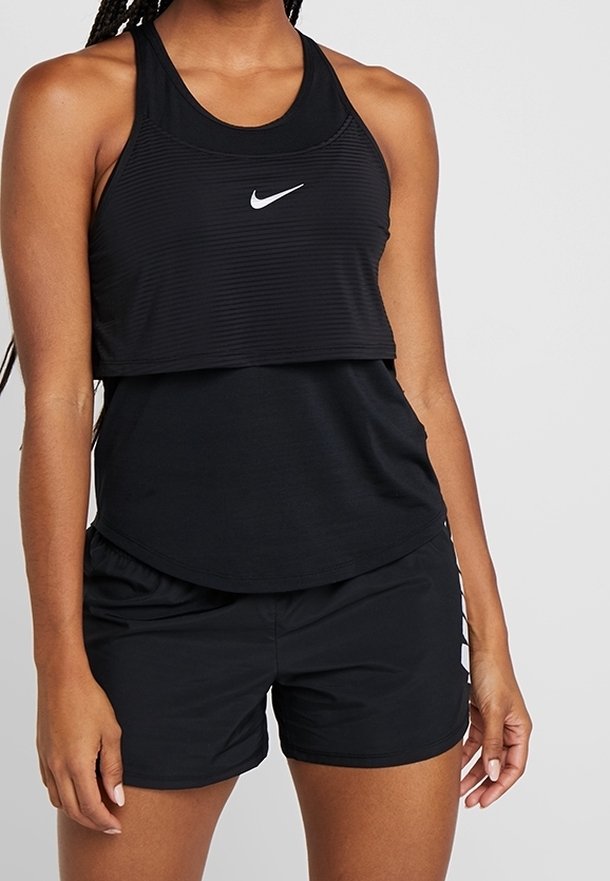 Femme portant un haut sans manches Nike noir superposé et un short de sport noir avec logo sur la cuisse gauche sur fond gris clair.