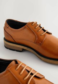 Chaussures en cuir marron avec une finition lisse, un bout arrondi et six œillets. La semelle présente un design superposé avec un bord texturé noir.