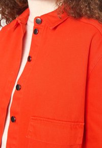 Detailní záběr na osobu, která má na sobě jasně oranžovou zapínací bundu přes svetr s vysokým límcem v světle krémové barvě a má kudrnaté vlasy.