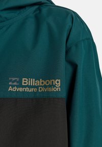 Tarn- und schwarze wasserdichte Jacke aus strukturiertem Material, mit Kapuze und goldenem Logo "Billabong Adventure Division" auf der Brust.