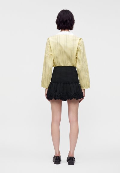 Personne aux cheveux courts et foncés, vue de dos, portant un chemisier jaune à fines rayures, une mini-jupe noire à volants et des chaussures noires sur fond blanc.