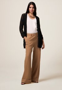 Cardigan nero a maglia aperta sopra una maglietta bianca abbinato a pantaloni marroni a vita alta e gamba larga. Collana chunky notevole e sandali con tacco.