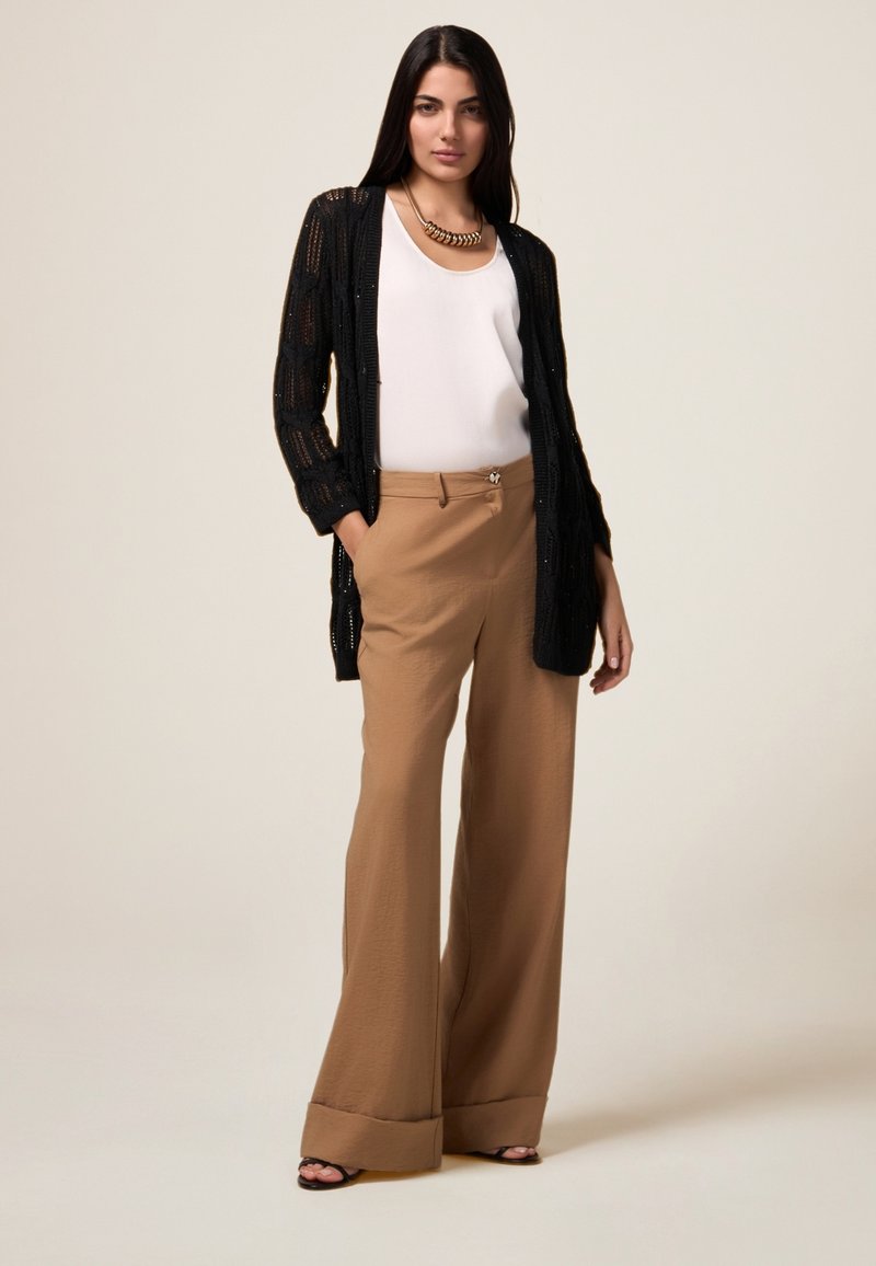Cardigan nero a maglia aperta sopra una maglietta bianca abbinato a pantaloni marroni a vita alta e gamba larga. Collana chunky notevole e sandali con tacco.