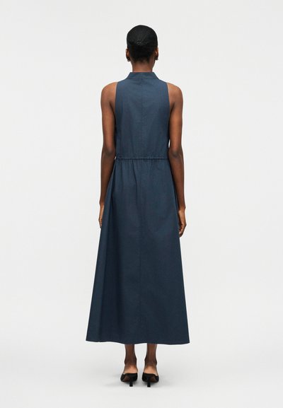 Γυναίκα που στέκεται με την πλάτη προς τον θεατή, φορώντας αμάνικο, σκουρομπλε φόρεμα μέχρι τους αστραγάλους και μαύρα παπούτσια με τακούνι σε απλό φόντο.