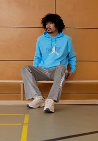 Modrá mikina s kapucňou s logom, sivé pruhované nohavice a béžové tenisky. Sedí na lavičke pred dreveným pozadím v telocvični.