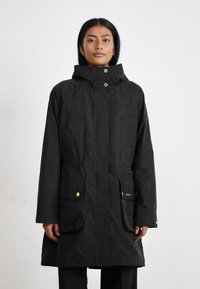 Zwarte waterdichte parka-jack met gouden hardware, voorzakken, verstelbare capuchon en elastische taille details. Gladde textuur en knielange ontwerp.