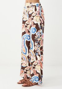 Weite Hosen in einem braunen Grundton mit einem floralen Muster, das blaue, rosa und cremefarbene Akzente aufweist. Weicher Stoff mit einem fließenden Design. Braune Sandalen.