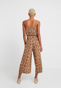 Vrouw met kort blond haar staat met haar rug naar ons gekeerd en draagt een bruine dierenprint wijde jumpsuit en beige blokhak sandalen.