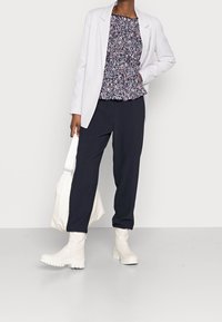 Blazer bianco sopra un top peplum floreale, pantaloni scuri e stivali alla caviglia bianchi. Modella tiene una borsa tote crema. Texture lisce e design contemporaneo.