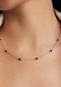Srebrny naszyjnik łańcuszkowy z okrągłymi czarnymi koralikami umieszczonymi w odstępach wzdłuż całej długości, spoczywający tuż nad obojczykiem.