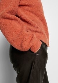 Pull en mohair orange à coupe large avec des poignets côtelés, associé à un pantalon en cuir marron avec une poche latérale et une texture lisse.